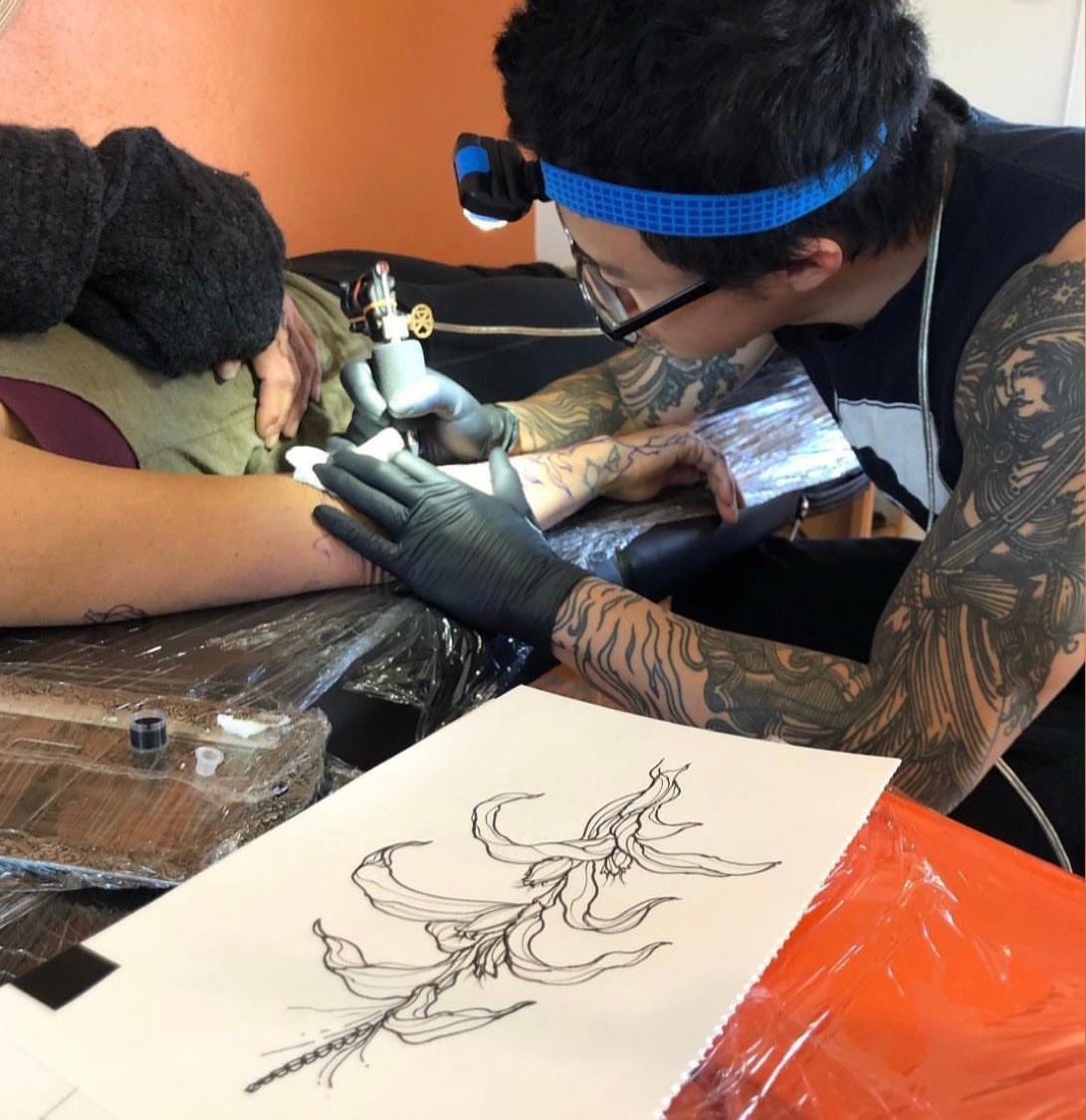 Jason working on my tattoo a man tattoos a corn plant onto an arm, the image is drawn on paper in the foreground 