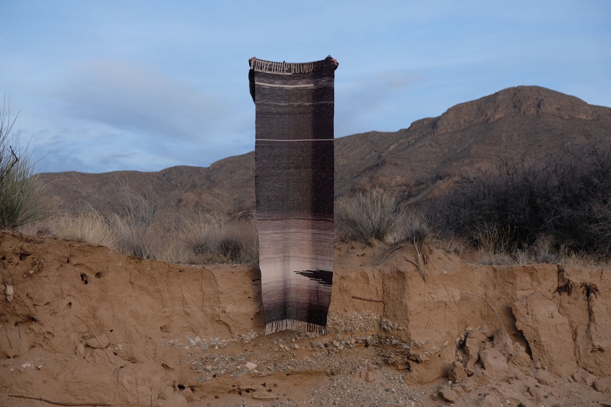 A shawl is held up on the banks of an arroyo, the person holding it obscured. The shawl is black, white with gray gradations. There is a mountain in the background. 