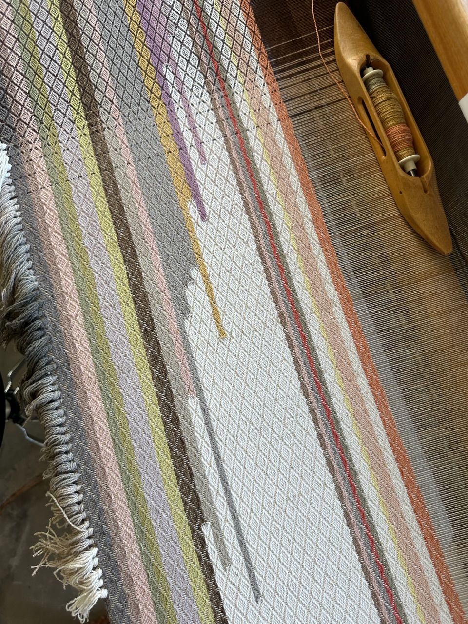 A detailed image of textured diamond pattern fabric on a loom, in soft naturally dyed shades of the rainbow.