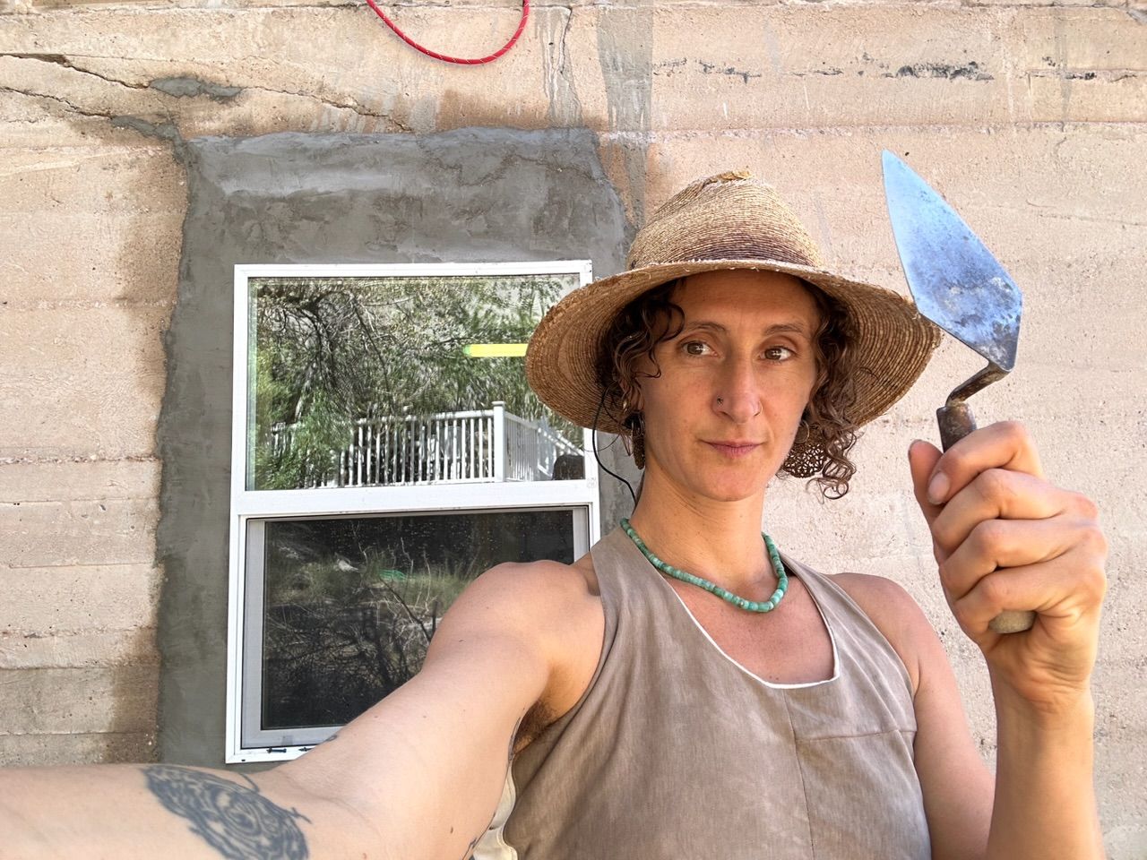 a woman in a cowboy hat and brown shirt holds a pointed trowel in front of a freshly stuccoed window