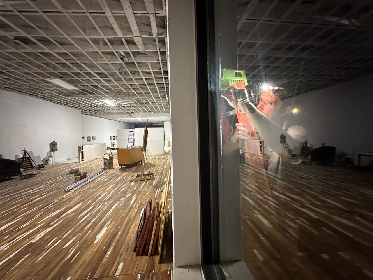 A slightly surreal image of a man on a ladder working on the outside of a window at night while the inside of a white walled, wood flowed room is well lit inside