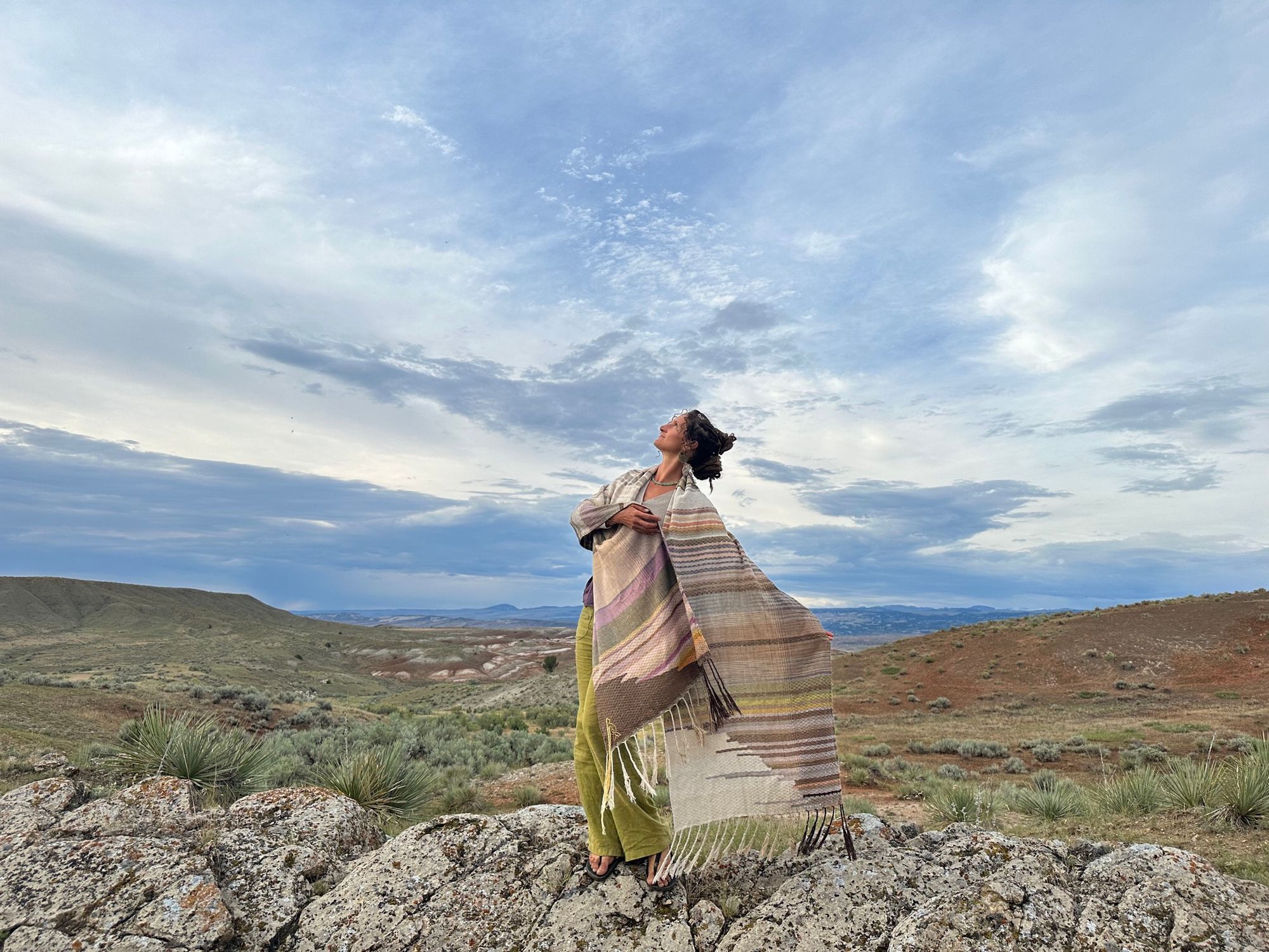 a woman wears a shawl of handwoven fabric of Naturally dyed subtle rainbow hues in a mountain landscape