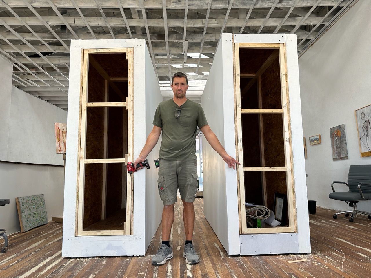 A man in a green shirt and shorts stands holding a red drill pointing to two door frames on while movable walls in a gallery space with a wood floor