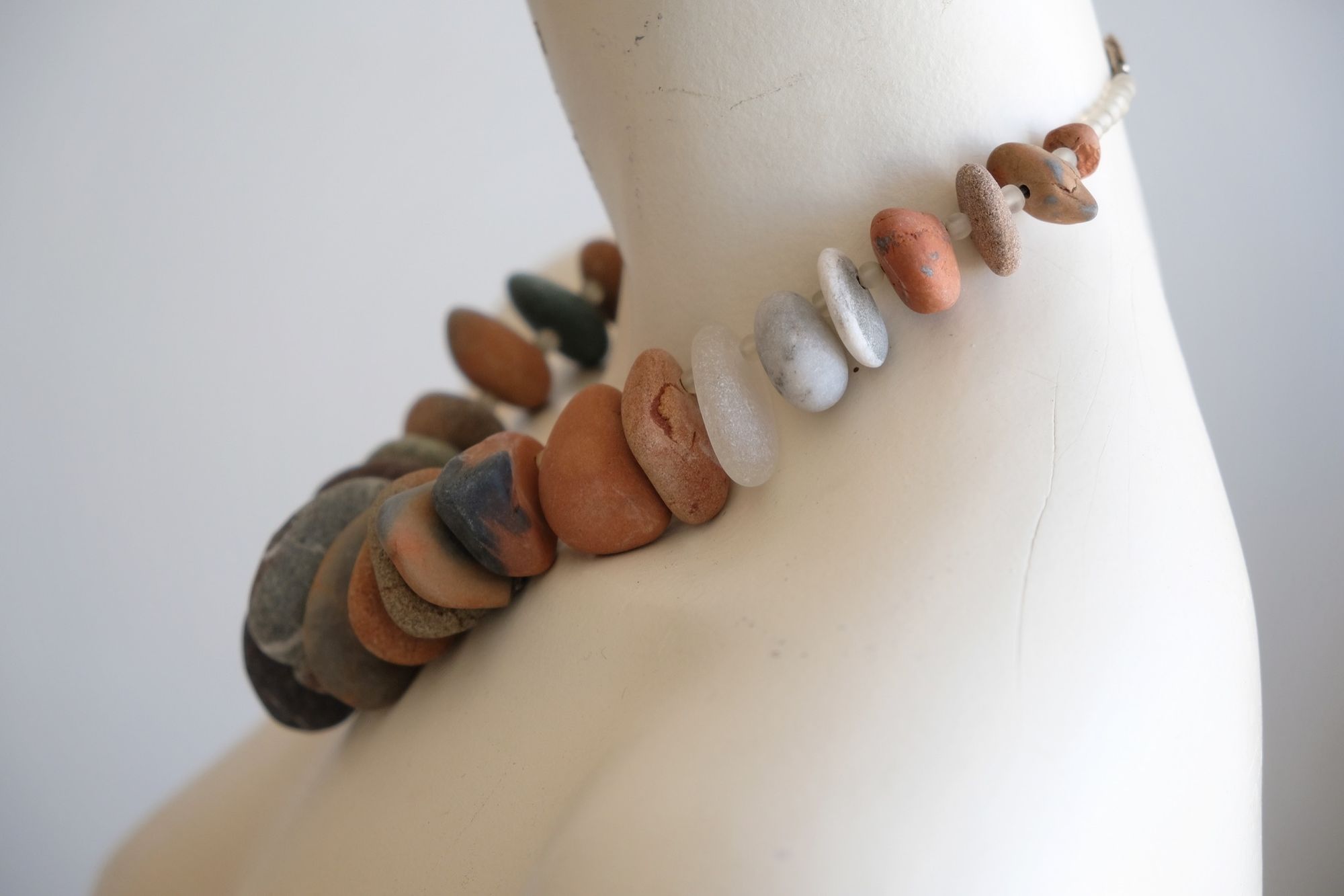 a white mannequin wears a necklace of smooth roundish ocean tumbled stones of brick red, grey and white