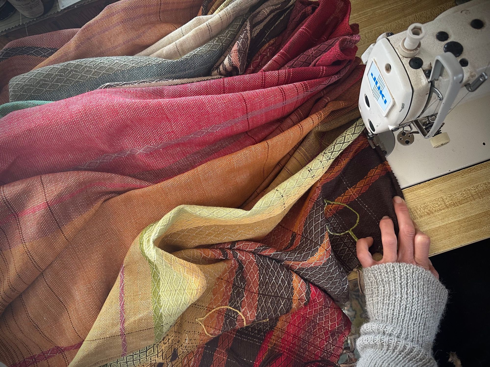a hand with a light grey sweater carefully holds the hem of a large piece of handwoven brown, red, orange and yellow piece of fabric being sewn on a Juki sewing machine