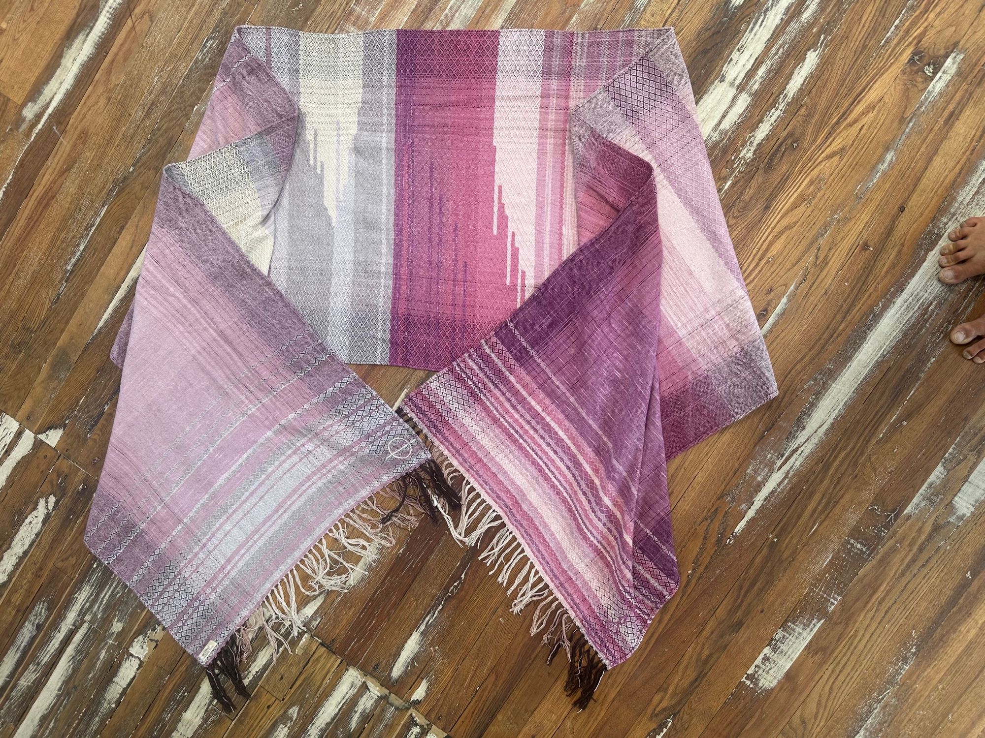 Handwoven fabric lays on a wood floor. The fabric is naturally dyed with cochineal in all shades of pink and fuchsia