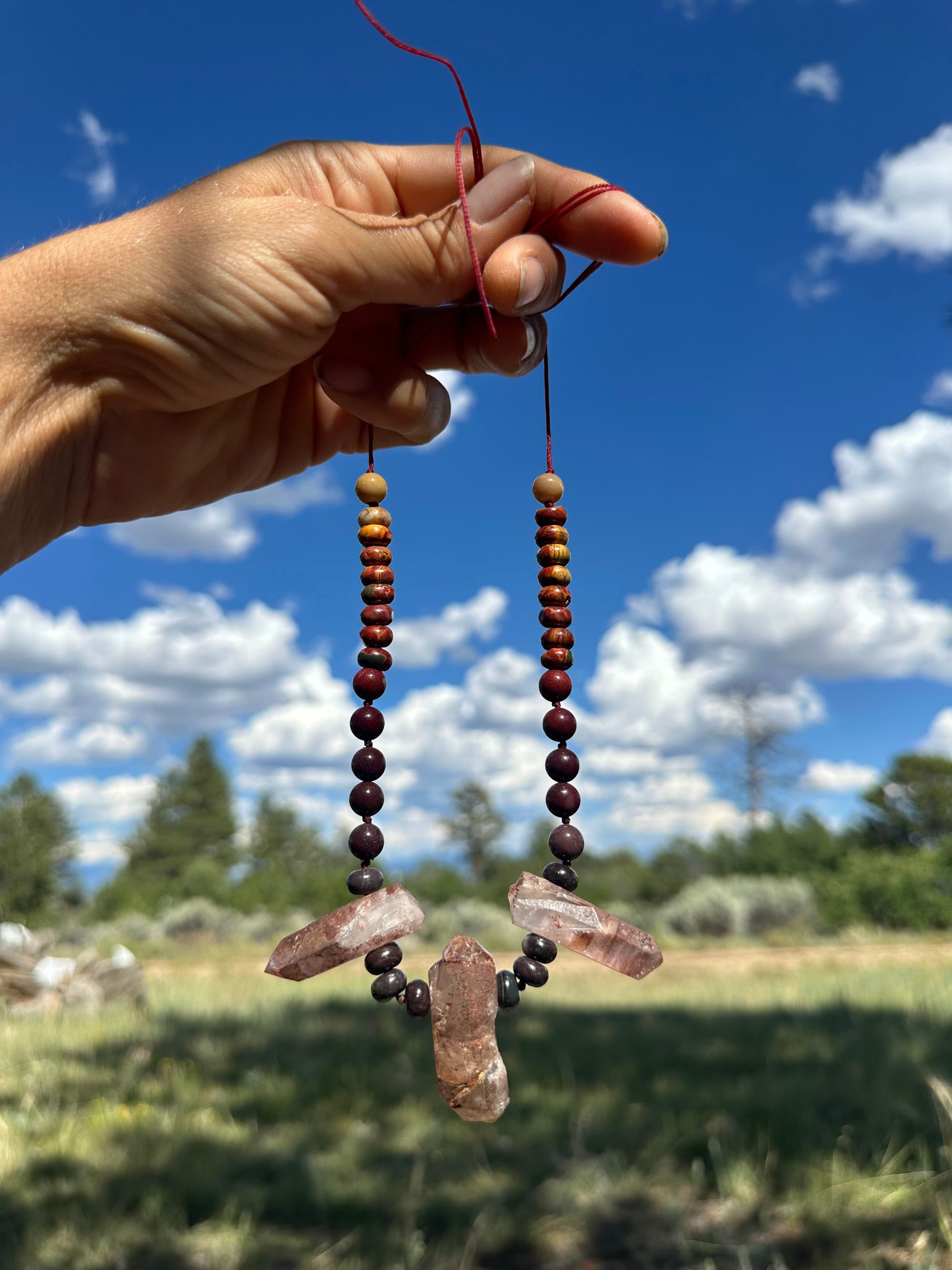 A handholds, a string of rainbow colored stone beads with three red quartz points against a bright blue sky and mountain Vista background