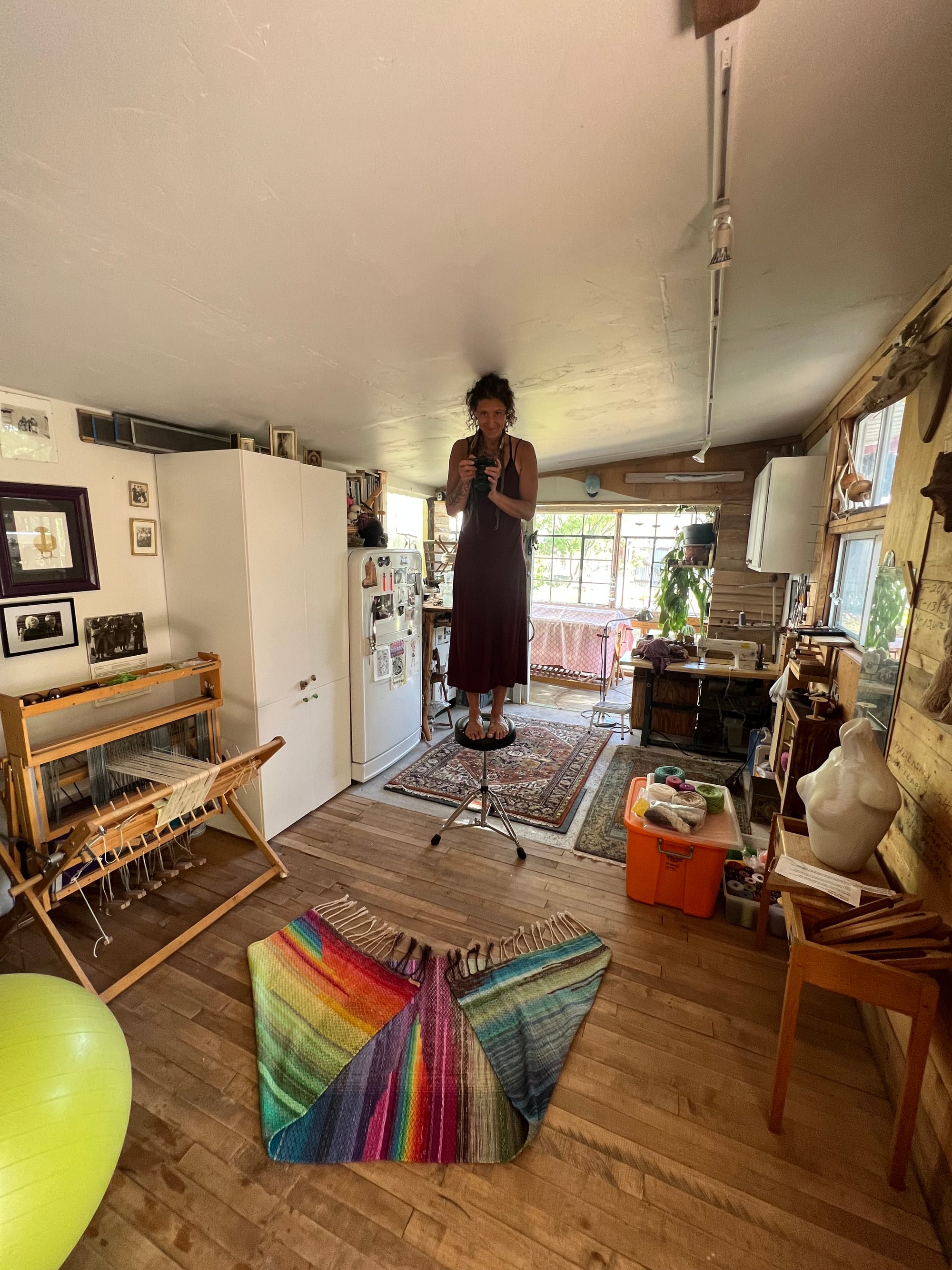 A woman in a black dress stands on a stool in a small studio room with weaving tools all around