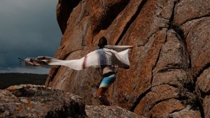A woman with a long shawl flips it into the wind while standing on lumpy, featured rocks.