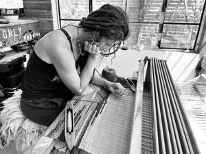 A black and white photo of a woman sits at a loom looking intently at the weaving and three shuttles sitting on the warp. 