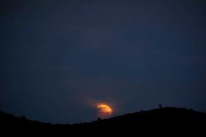 A deep red orange moon rises behind thick grey smoke and the silhouette of a mountain 