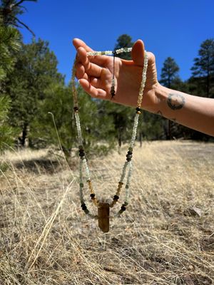 a hand holds a green peridot and deep yellow citrine necklace in front of green pine trees and yellow grass. 