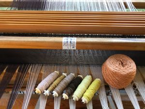 six bobbins of raw silk and a ball of salmon colored yarn sit on the white and brown strings of a loom