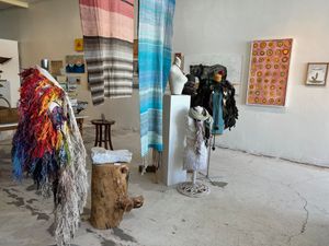 Two cloaks are installed on manequins with weavings hanging from the ceiling and paintings on the wall.