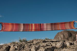 a long length of handwoven fabric in deep reds, purple, pink and orange is stretched across a blue sky 