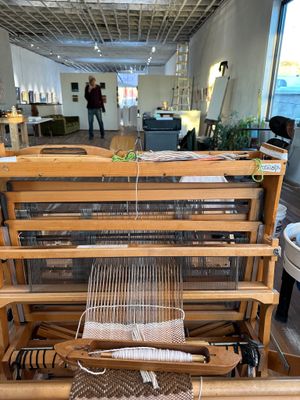 a wooden loom with an earth tone scarf being woven on it, in a painted white gallery-studio space