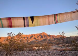 A long piece of handwoven fabric, in many colorful shades, is stretched against a sunset lit desert mountain