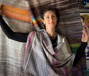 a woman stands in front of of, and is wrapped in a length of handwoven fabric, in earth tones and rainbows