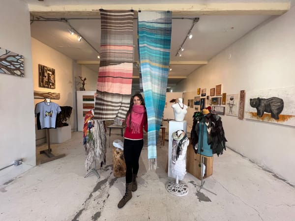 A woman stands in the middle of the frame between two handwoven textiles hanging from the celing. 