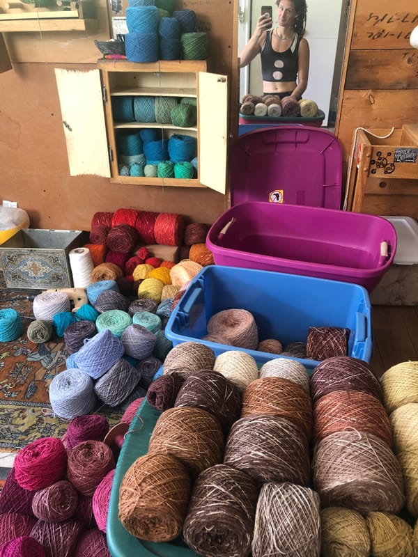 a rainbow palette of balls of think yarn sits in a studio setting, a woman is seen reflected in a mirror