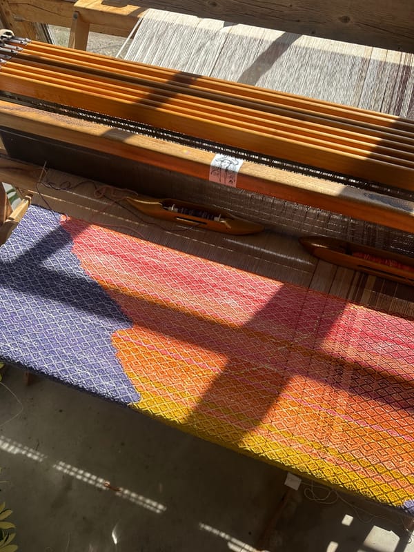 a large loom is brightly illuminated by windows, the weaving on it is bright sunrise colors and purple